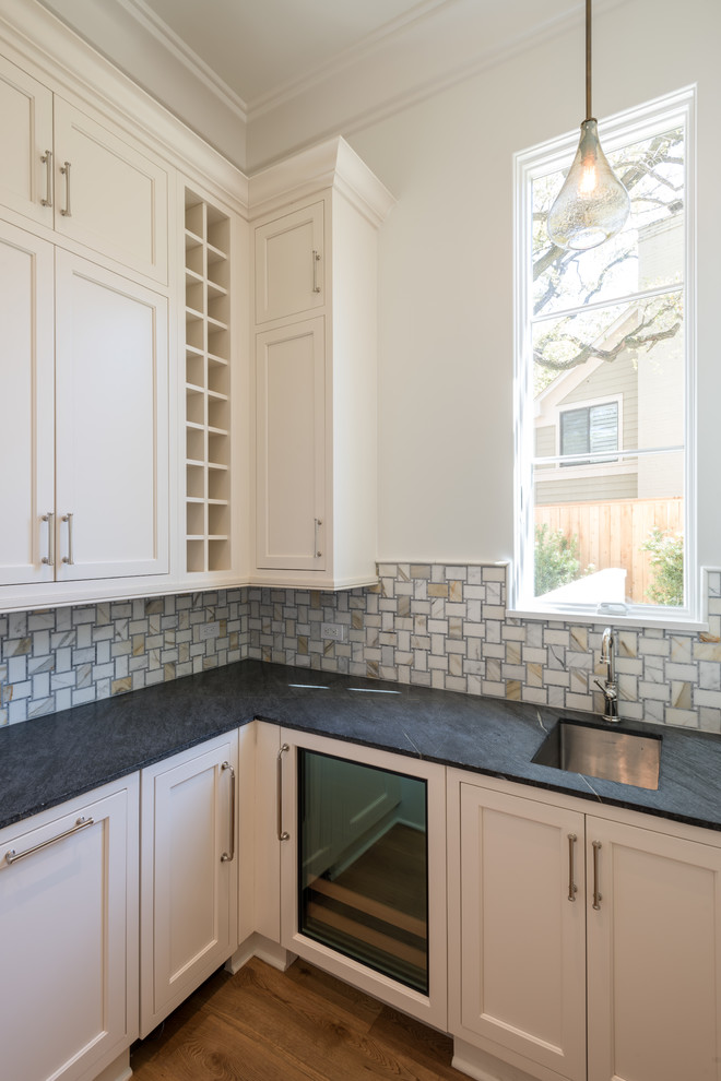 Inspiration for a small transitional l-shaped wet bar in Dallas with an undermount sink, beaded inset cabinets, beige cabinets, soapstone benchtops, beige splashback, mosaic tile splashback and light hardwood floors.