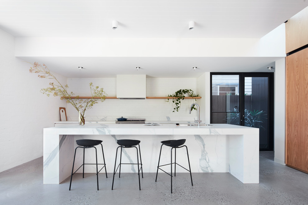Large contemporary galley open plan kitchen in Melbourne with an undermount sink, medium wood cabinets, marble benchtops, white splashback, brick splashback, stainless steel appliances, concrete floors, with island, grey floor and white benchtop.
