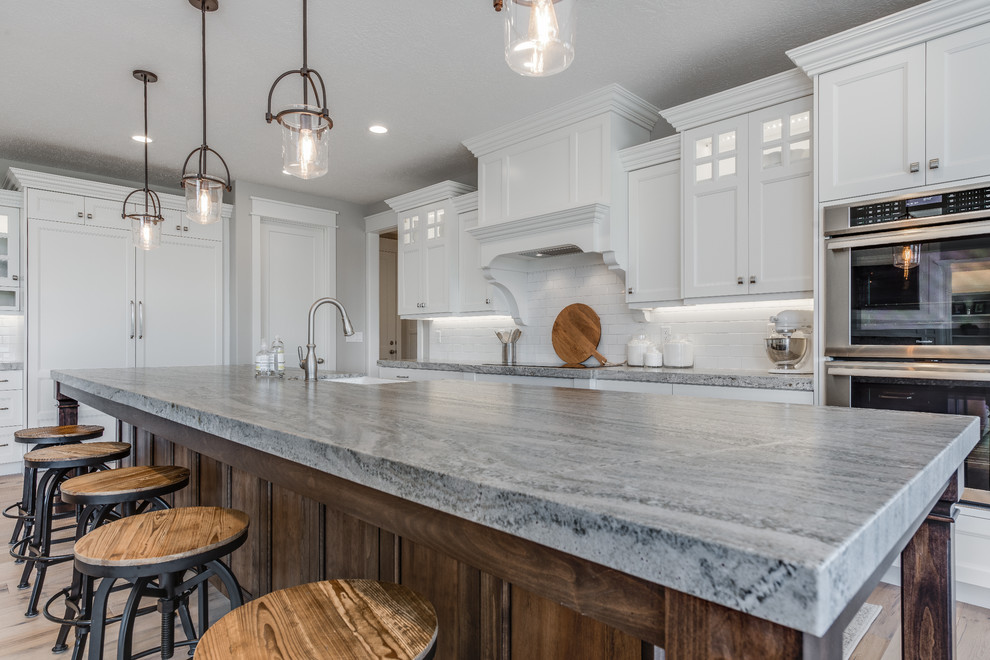 Inspiration for a large arts and crafts l-shaped eat-in kitchen in Salt Lake City with a farmhouse sink, recessed-panel cabinets, white cabinets, white splashback, brick splashback, stainless steel appliances, light hardwood floors, with island and brown floor.