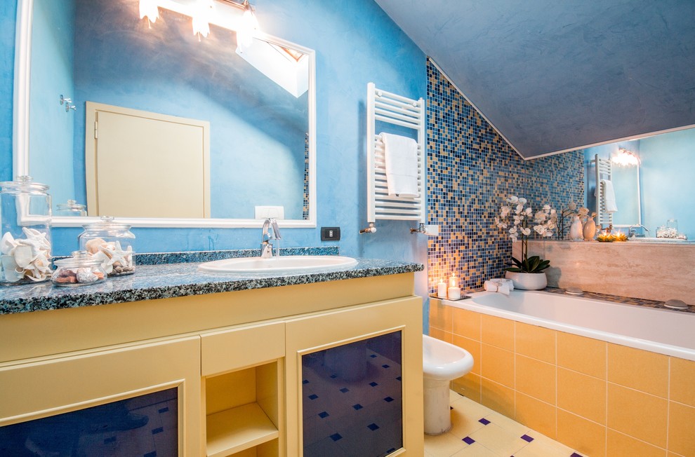Beach style 3/4 bathroom in Rome with shaker cabinets, yellow cabinets, an alcove tub, blue tile, yellow tile, mosaic tile, blue walls, a drop-in sink, yellow floor and black benchtops.