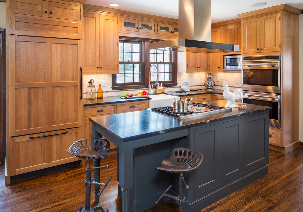 Inspiration for a mid-sized country l-shaped open plan kitchen in Minneapolis with a farmhouse sink, flat-panel cabinets, soapstone benchtops, white splashback, subway tile splashback, with island, medium wood cabinets, stainless steel appliances, dark hardwood floors and brown floor.