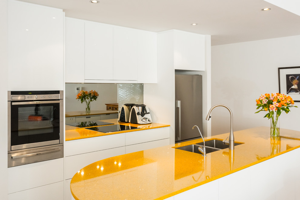 Photo of a small contemporary galley open plan kitchen in Perth with an undermount sink, flat-panel cabinets, white cabinets, glass benchtops, metallic splashback, glass sheet splashback, stainless steel appliances and with island.