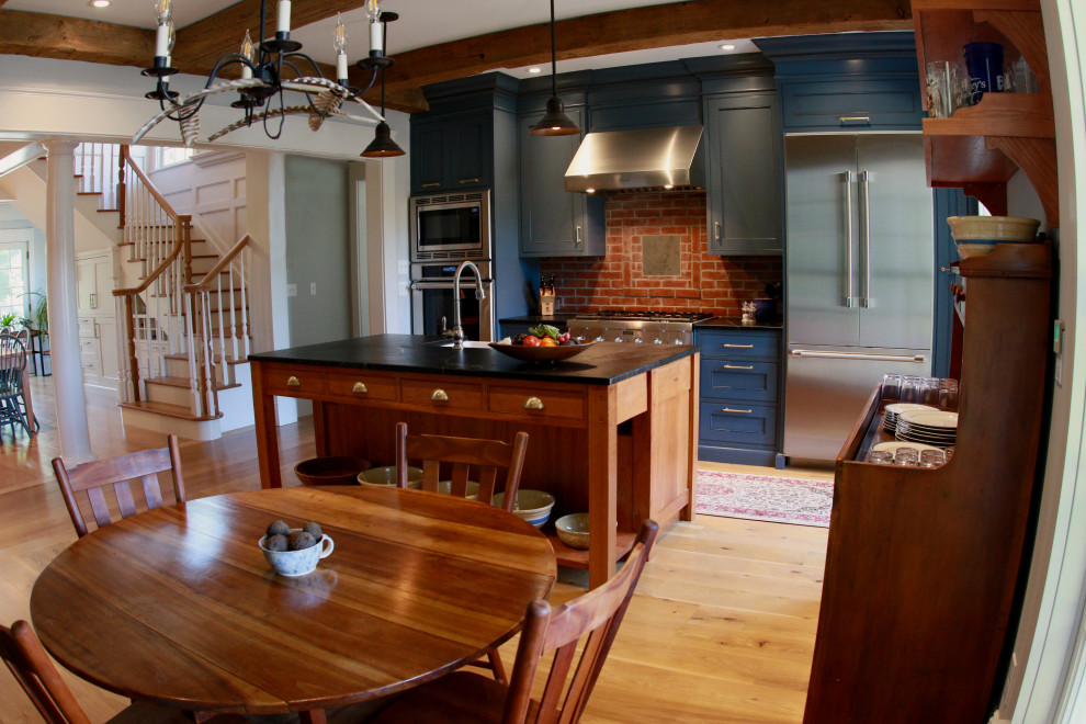 Photo of a mid-sized country single-wall eat-in kitchen in Boston with a farmhouse sink, shaker cabinets, blue cabinets, soapstone benchtops, multi-coloured splashback, brick splashback, stainless steel appliances, medium hardwood floors, with island, black benchtop and exposed beam.