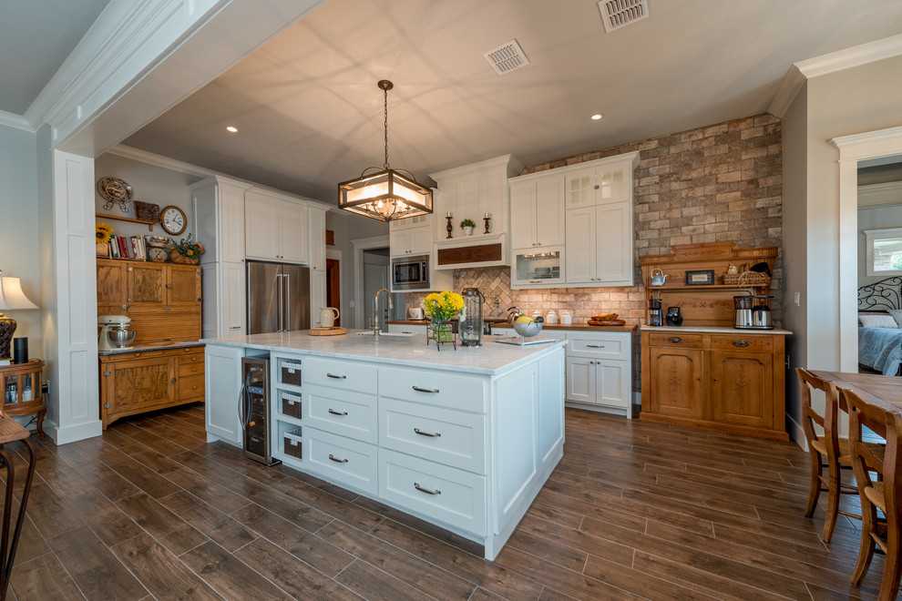 This is an example of a mid-sized arts and crafts l-shaped eat-in kitchen in Other with flat-panel cabinets, white cabinets, granite benchtops, brown splashback, stone tile splashback, stainless steel appliances, ceramic floors, with island, brown floor and a farmhouse sink.