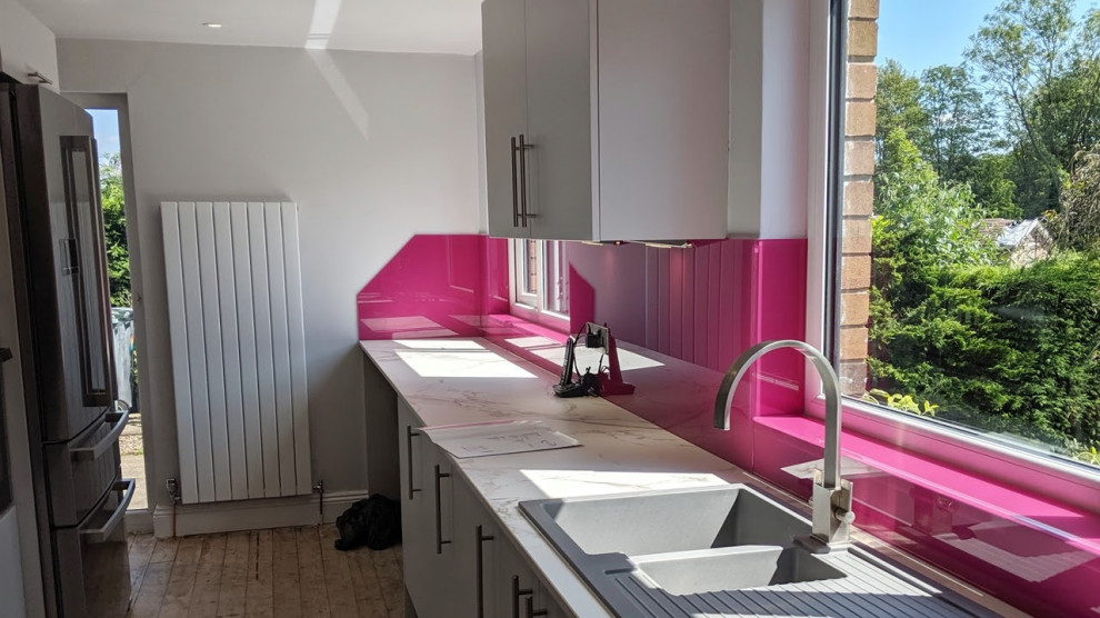 Inspiration for a mid-sized contemporary l-shaped separate kitchen in Manchester with a double-bowl sink, flat-panel cabinets, white cabinets, solid surface benchtops, pink splashback, glass sheet splashback, stainless steel appliances, laminate floors, brown floor and grey benchtop.