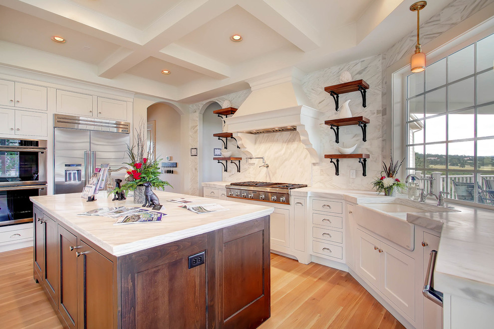This is an example of a transitional galley kitchen in Denver with a farmhouse sink, shaker cabinets, white cabinets, grey splashback, stainless steel appliances, light hardwood floors and multiple islands.