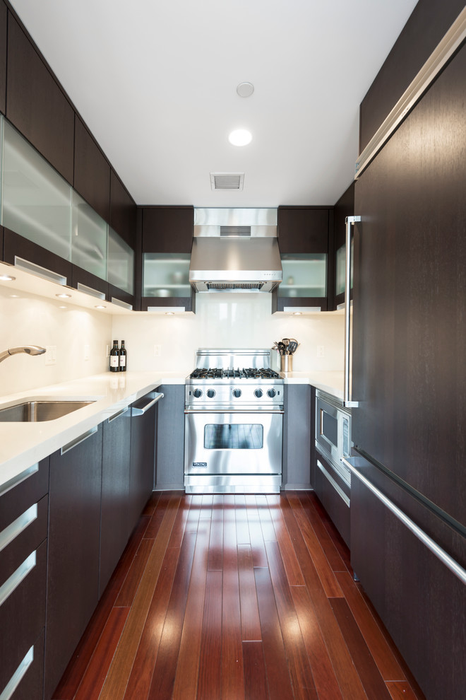 Photo of a small modern galley separate kitchen in New York with a drop-in sink, glass-front cabinets, brown cabinets, limestone benchtops, white splashback, stone tile splashback, stainless steel appliances, dark hardwood floors and no island.