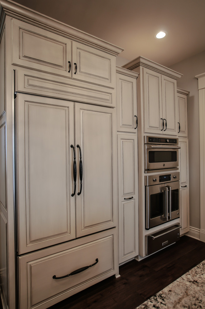 Inspiration for a large traditional l-shaped eat-in kitchen in Omaha with a farmhouse sink, raised-panel cabinets, distressed cabinets, granite benchtops, multi-coloured splashback, stone tile splashback, panelled appliances, dark hardwood floors, with island, brown floor and beige benchtop.