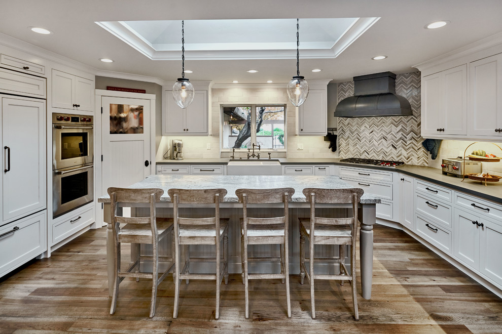 Large country u-shaped open plan kitchen in San Francisco with a farmhouse sink, beaded inset cabinets, white cabinets, granite benchtops, grey splashback, stone tile splashback, panelled appliances, light hardwood floors and with island.