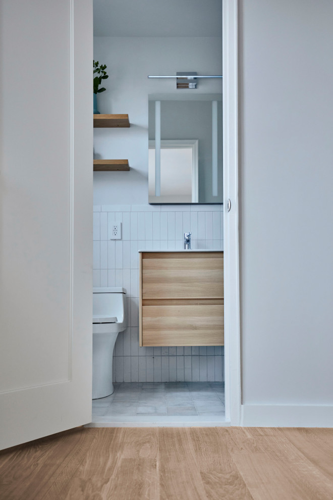 Small contemporary ensuite bathroom in New York with flat-panel cabinets, light wood cabinets, an alcove shower, a one-piece toilet, white tiles, ceramic tiles, grey walls, an integrated sink, solid surface worktops, grey floors, a sliding door, white worktops, a wall niche, a single sink, a floating vanity unit, a drop ceiling and marble flooring.