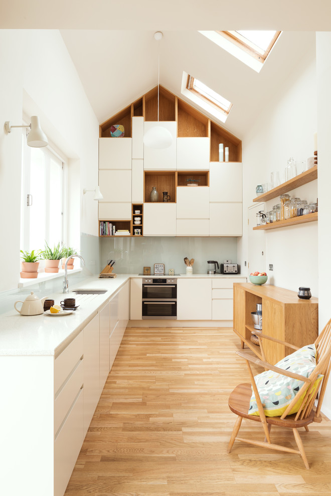 Design ideas for a mid-sized contemporary l-shaped kitchen in London with flat-panel cabinets, white cabinets, recycled glass benchtops, blue splashback, glass sheet splashback, no island, white benchtop, an undermount sink, light hardwood floors and beige floor.