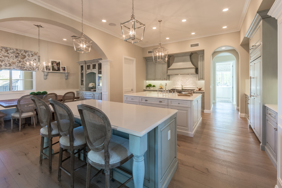 Example of a mid-sized transitional u-shaped medium tone wood floor and brown floor open concept kitchen design in Phoenix with an undermount sink, raised-panel cabinets, gray cabinets, solid surface countertops, white backsplash, ceramic backsplash, stainless steel appliances, two islands and white countertops