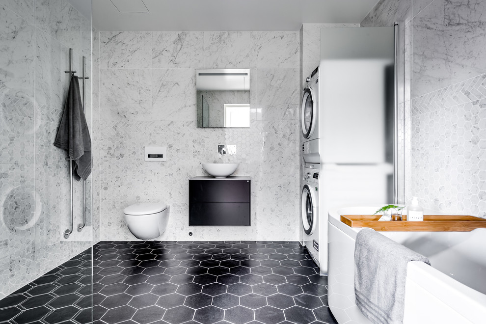 This is an example of a large contemporary bathroom in Stockholm with black cabinets, a freestanding tub, a wall-mount toilet, white tile, marble, a vessel sink, black floor and flat-panel cabinets.