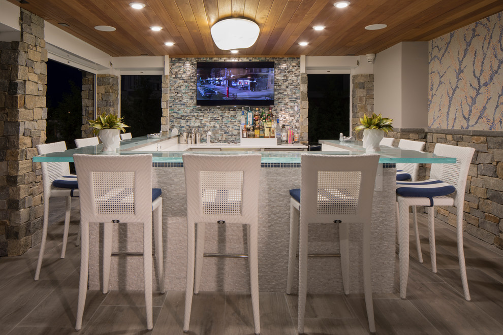 This is an example of a mid-sized beach style u-shaped seated home bar in Philadelphia with glass benchtops, multi-coloured splashback, mosaic tile splashback, blue benchtop, medium hardwood floors and brown floor.