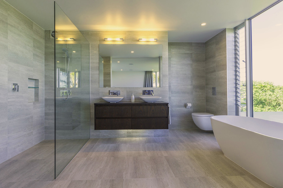 Design ideas for a mid-sized contemporary master bathroom in Auckland with dark wood cabinets, a freestanding tub, a curbless shower, a wall-mount toilet, gray tile, grey walls, ceramic floors and wood benchtops.