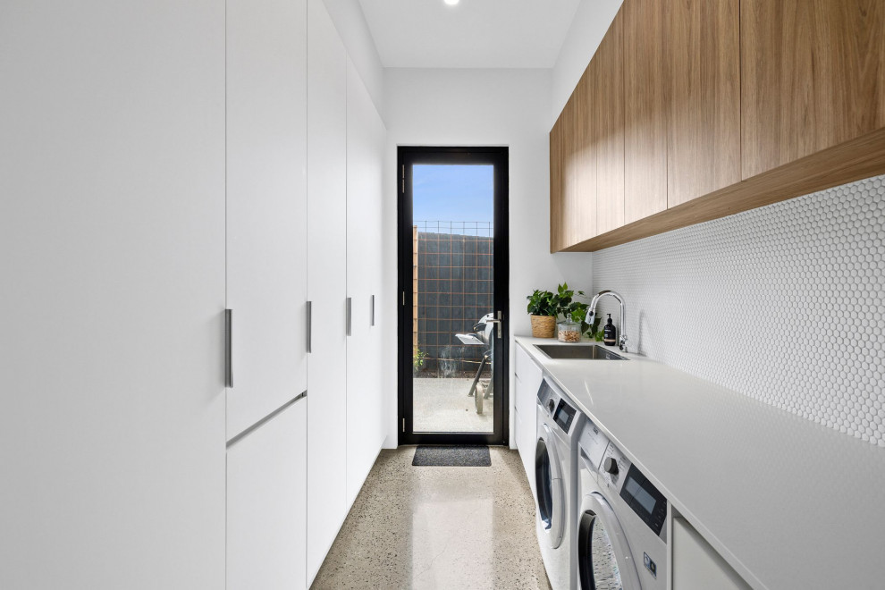 Inspiration for a large contemporary galley dedicated laundry room in Geelong with a drop-in sink, flat-panel cabinets, white cabinets, quartz benchtops, white splashback, mosaic tile splashback, white walls, concrete floors, a side-by-side washer and dryer and white benchtop.