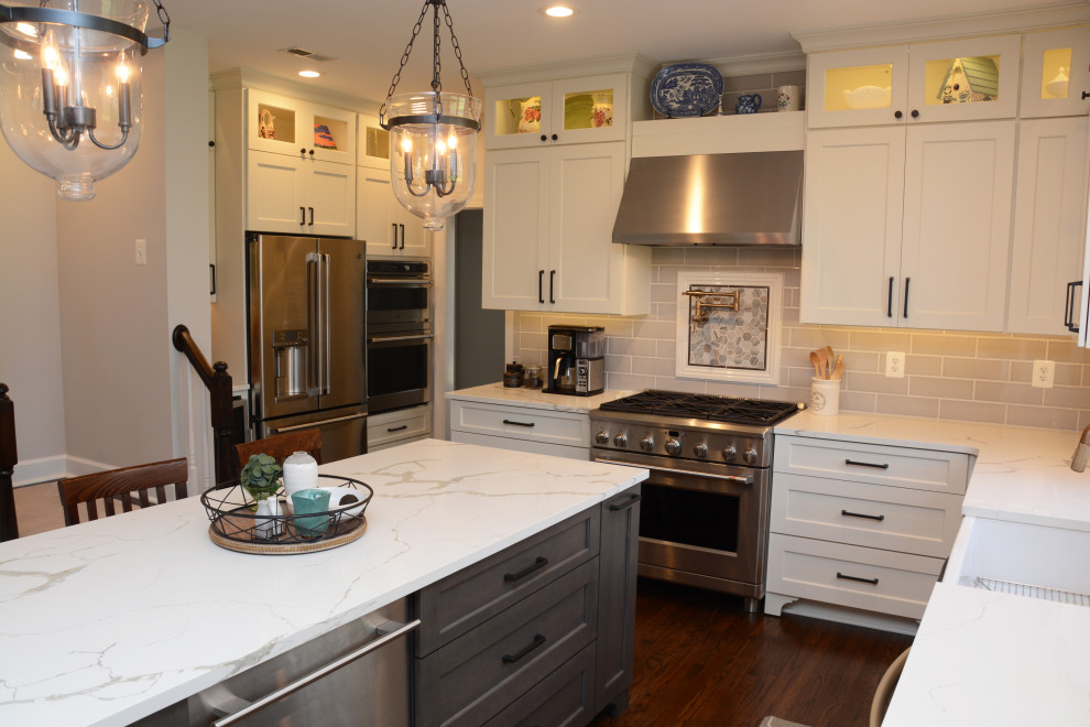 Photo of a large traditional l-shaped eat-in kitchen in DC Metro with a farmhouse sink, recessed-panel cabinets, white cabinets, quartz benchtops, grey splashback, stainless steel appliances, dark hardwood floors, with island, brown floor and white benchtop.