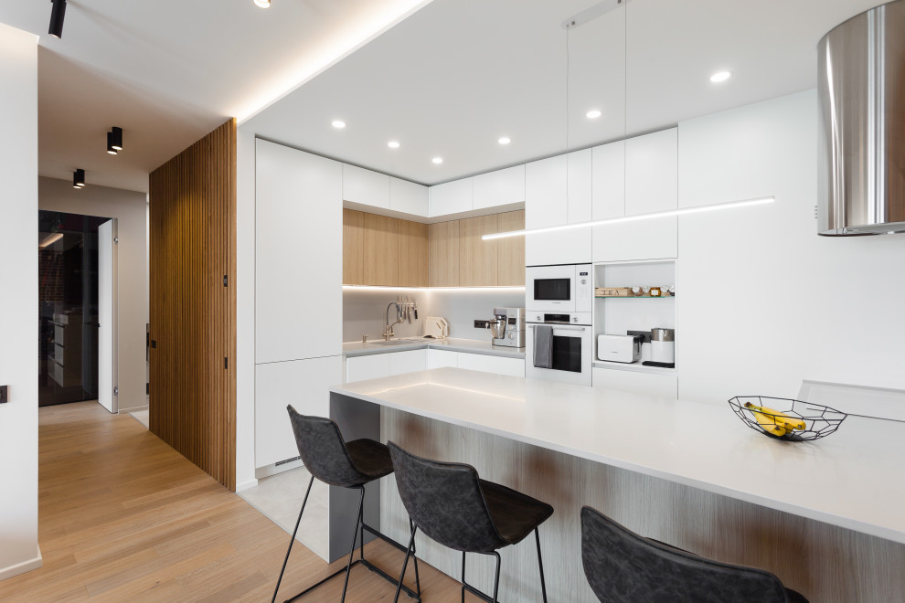 This is an example of a mid-sized contemporary l-shaped open plan kitchen in Moscow with an undermount sink, flat-panel cabinets, white cabinets, solid surface benchtops, grey splashback, white appliances, porcelain floors, a peninsula, grey floor, grey benchtop and recessed.
