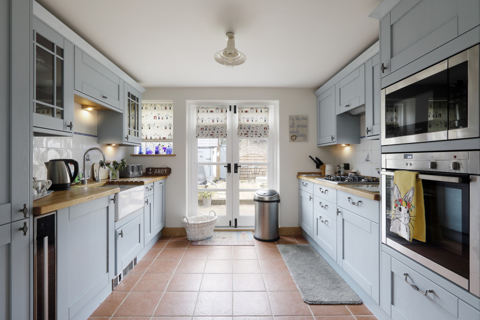 Design ideas for a mid-sized country galley separate kitchen in Sussex with a farmhouse sink, recessed-panel cabinets, blue cabinets, wood benchtops, white splashback, ceramic splashback, stainless steel appliances, terra-cotta floors, no island, red floor and brown benchtop.