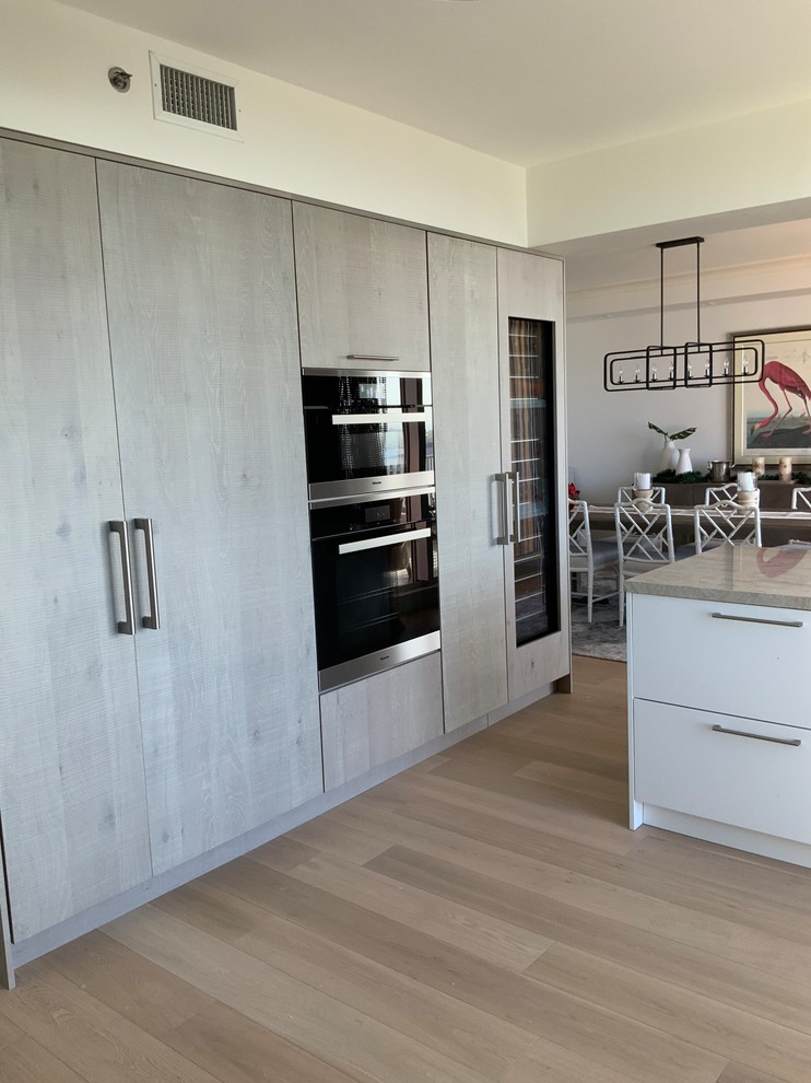 Photo of a mid-sized contemporary l-shaped kitchen in Chicago with a double-bowl sink, flat-panel cabinets, distressed cabinets, quartzite benchtops, grey splashback, stone slab splashback, stainless steel appliances, light hardwood floors, with island, beige floor and grey benchtop.