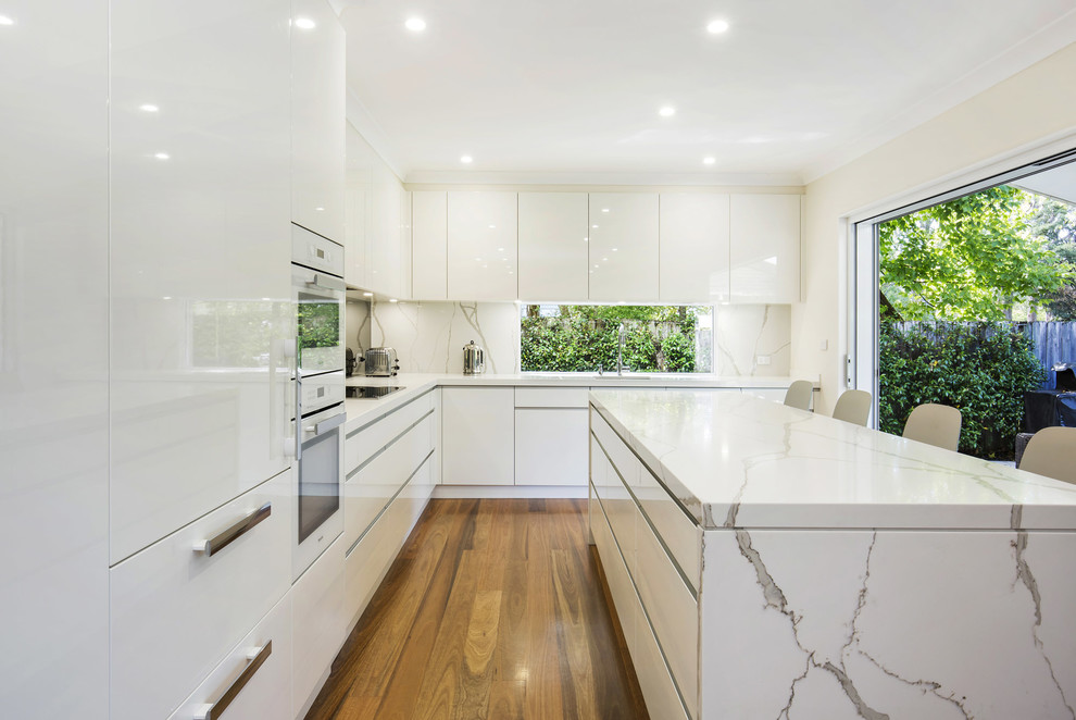 This is an example of a mid-sized contemporary l-shaped eat-in kitchen in Sydney with an undermount sink, flat-panel cabinets, white cabinets, quartz benchtops, white appliances, with island, multi-coloured splashback, window splashback and light hardwood floors.