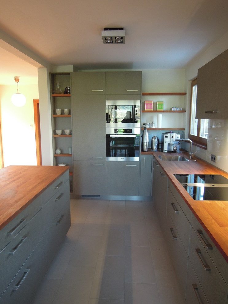 Mid-sized contemporary galley open plan kitchen in Frankfurt with a drop-in sink, flat-panel cabinets, grey cabinets, wood benchtops, white splashback, glass sheet splashback, stainless steel appliances, with island and light hardwood floors.