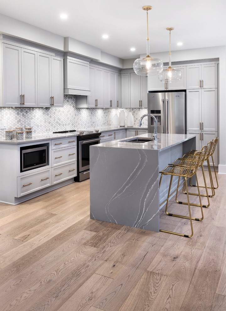 Transitional l-shaped kitchen in Ottawa with an undermount sink, recessed-panel cabinets, grey cabinets, quartz benchtops, grey splashback, mosaic tile splashback, stainless steel appliances, light hardwood floors, with island, brown floor and grey benchtop.