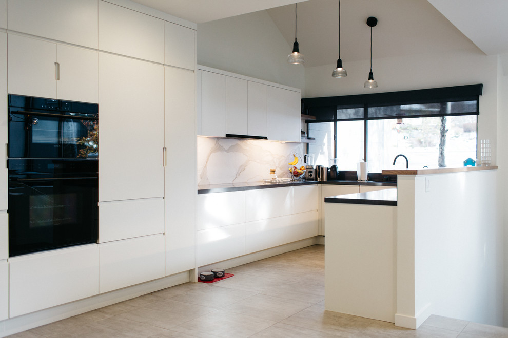 This is an example of a mid-sized modern u-shaped open plan kitchen in Salt Lake City with flat-panel cabinets, white cabinets, solid surface benchtops, white splashback, stone slab splashback, black appliances, ceramic floors and with island.