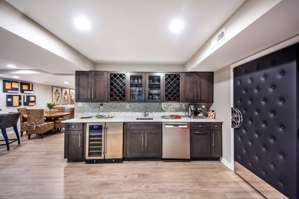 Mid-sized transitional single-wall wet bar in DC Metro with an undermount sink, shaker cabinets, brown cabinets, granite benchtops, white splashback, mosaic tile splashback, vinyl floors, beige floor and white benchtop.