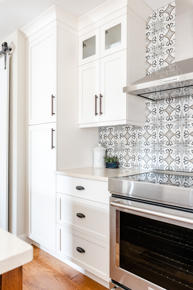 Mid-sized transitional l-shaped medium tone wood floor and brown floor kitchen pantry photo with an undermount sink, shaker cabinets, white cabinets, quartzite countertops, gray backsplash, porcelain backsplash, stainless steel appliances, an island and white countertops