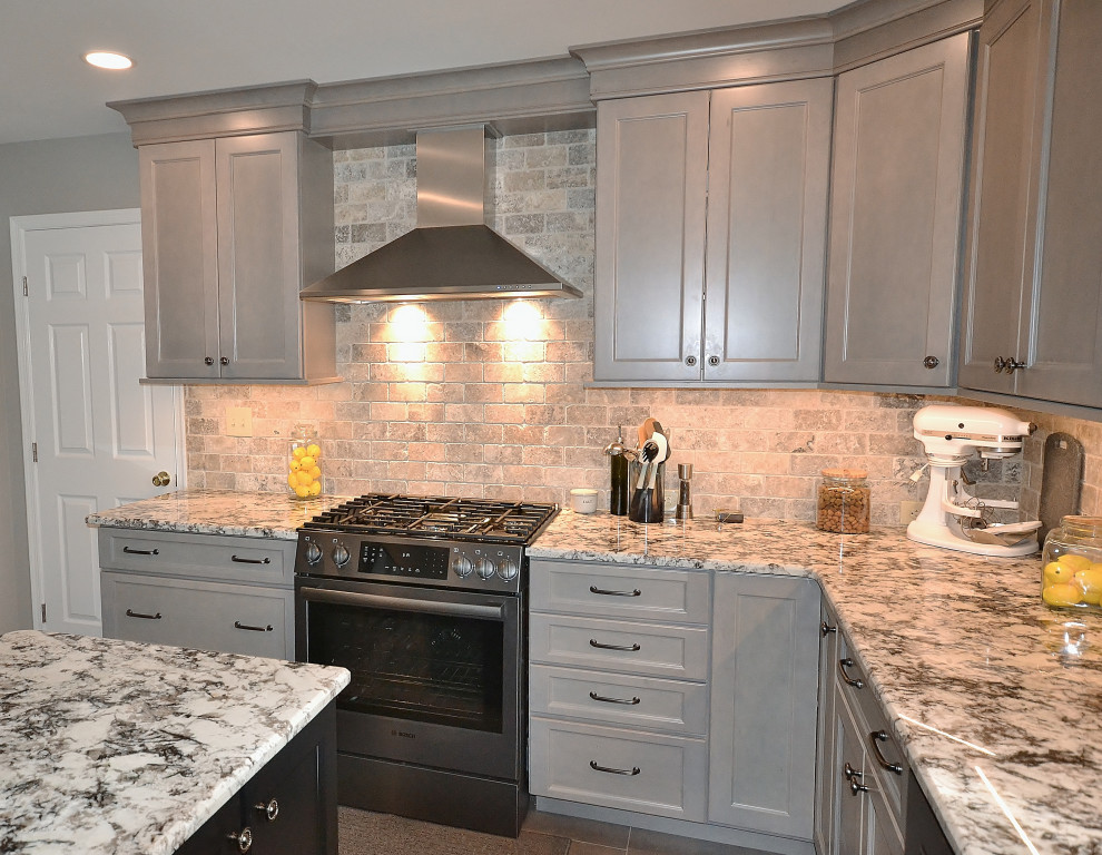 Photo of a large transitional l-shaped eat-in kitchen in Philadelphia with a farmhouse sink, raised-panel cabinets, grey cabinets, granite benchtops, beige splashback, travertine splashback, stainless steel appliances, vinyl floors, with island, beige floor and multi-coloured benchtop.