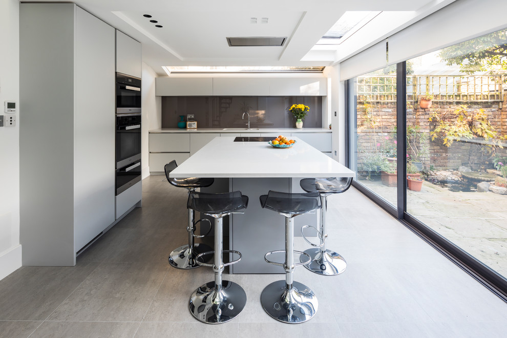 This is an example of a contemporary kitchen in London with flat-panel cabinets, grey cabinets, grey splashback, glass sheet splashback, black appliances, with island and grey floor.