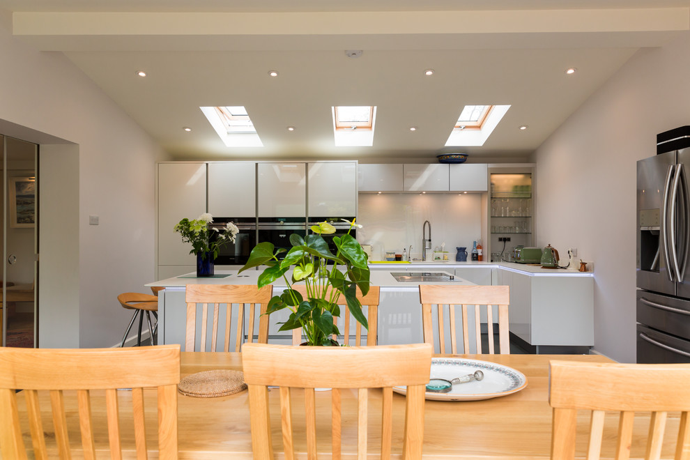 This is an example of a large contemporary single-wall open plan kitchen in Hampshire with an integrated sink, flat-panel cabinets, white cabinets, solid surface benchtops, white splashback, glass sheet splashback, stainless steel appliances, ceramic floors and with island.