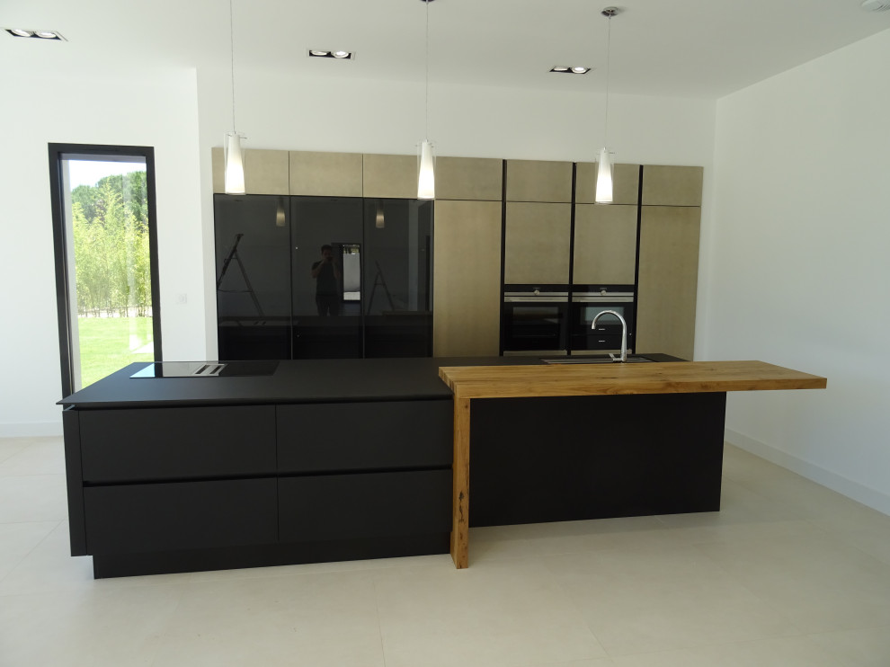 This is an example of a large modern galley open plan kitchen in Marseille with an integrated sink, beaded inset cabinets, black cabinets, black appliances, ceramic floors, with island, beige floor and black benchtop.
