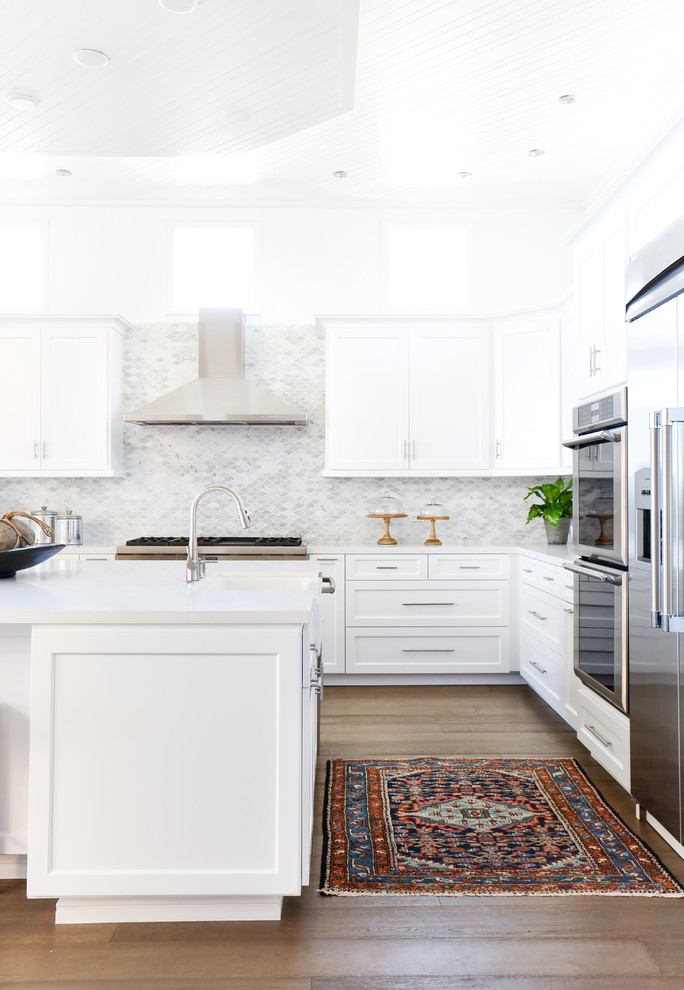 This is an example of a large beach style l-shaped open plan kitchen in Orange County with a drop-in sink, shaker cabinets, white cabinets, quartz benchtops, grey splashback, mosaic tile splashback, stainless steel appliances, dark hardwood floors and with island.