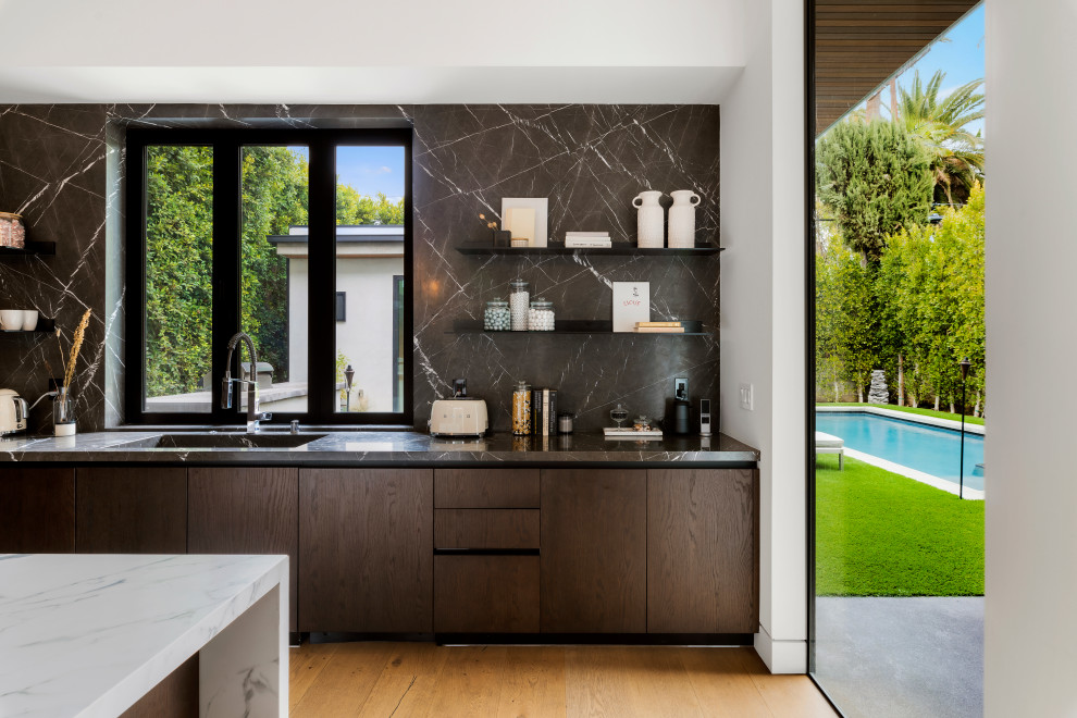 Photo of a mid-sized contemporary galley eat-in kitchen in Los Angeles with flat-panel cabinets, dark wood cabinets, marble benchtops, black splashback, an integrated sink, light hardwood floors, beige floor, marble splashback, with island and black benchtop.