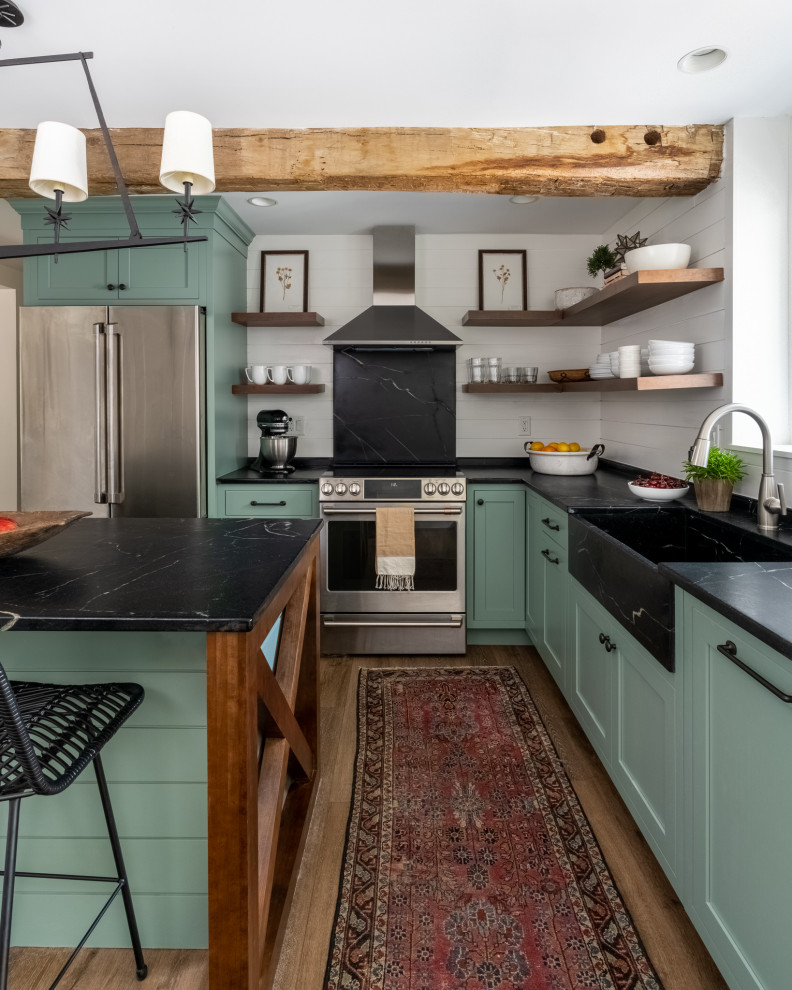Photo of a small country l-shaped separate kitchen in Philadelphia with a farmhouse sink, shaker cabinets, green cabinets, soapstone benchtops, black splashback, stone slab splashback, stainless steel appliances, laminate floors, with island, brown floor and black benchtop.