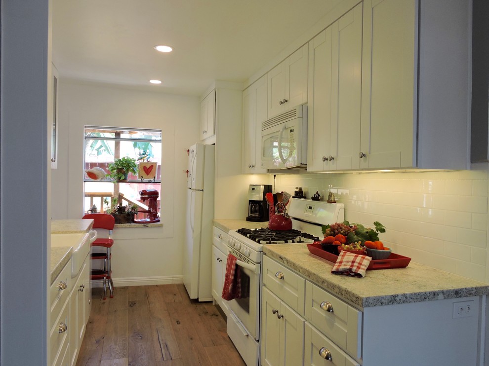 This is an example of a small beach style galley kitchen in San Diego with a farmhouse sink, shaker cabinets, white cabinets, granite benchtops, white splashback, porcelain splashback, white appliances, medium hardwood floors and no island.