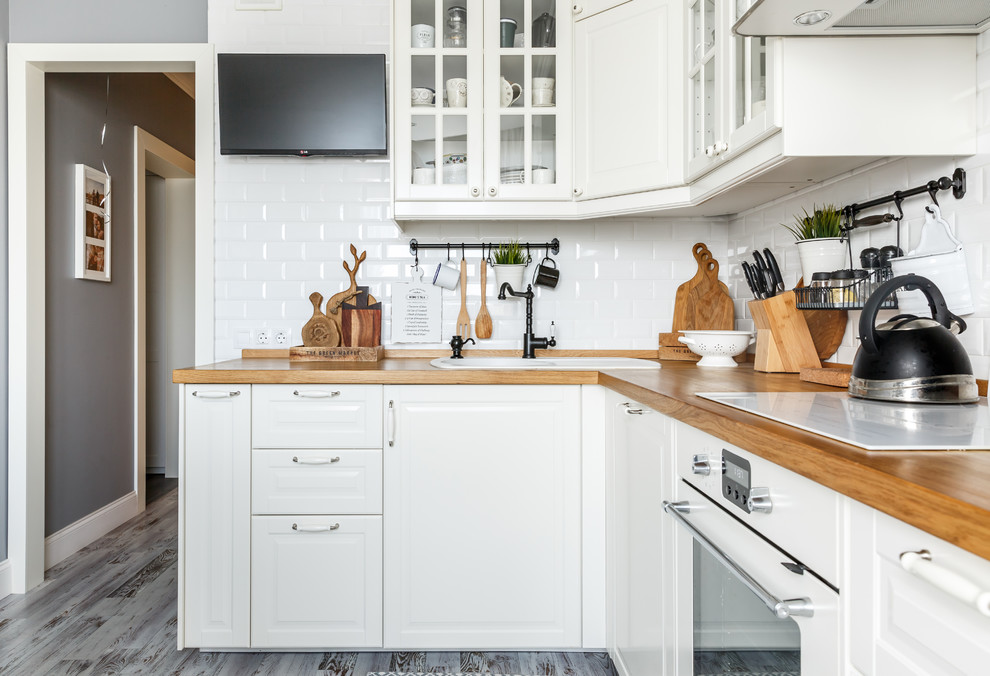 This is an example of a scandinavian l-shaped eat-in kitchen in Moscow with a drop-in sink, raised-panel cabinets, white cabinets, wood benchtops, white splashback, subway tile splashback, white appliances, dark hardwood floors, no island, grey floor and brown benchtop.