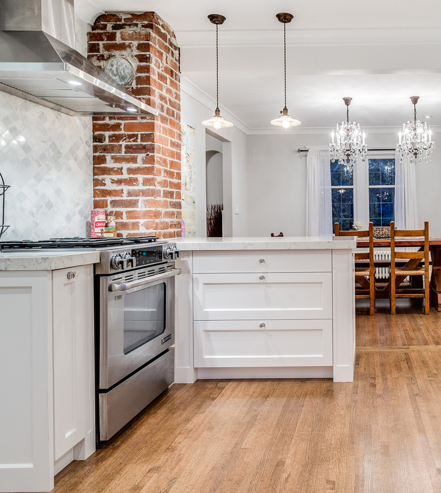 Inspiration for a mid-sized transitional l-shaped medium tone wood floor eat-in kitchen remodel in Vancouver with a farmhouse sink, shaker cabinets, white cabinets, quartzite countertops, white backsplash, stone tile backsplash, stainless steel appliances and a peninsula