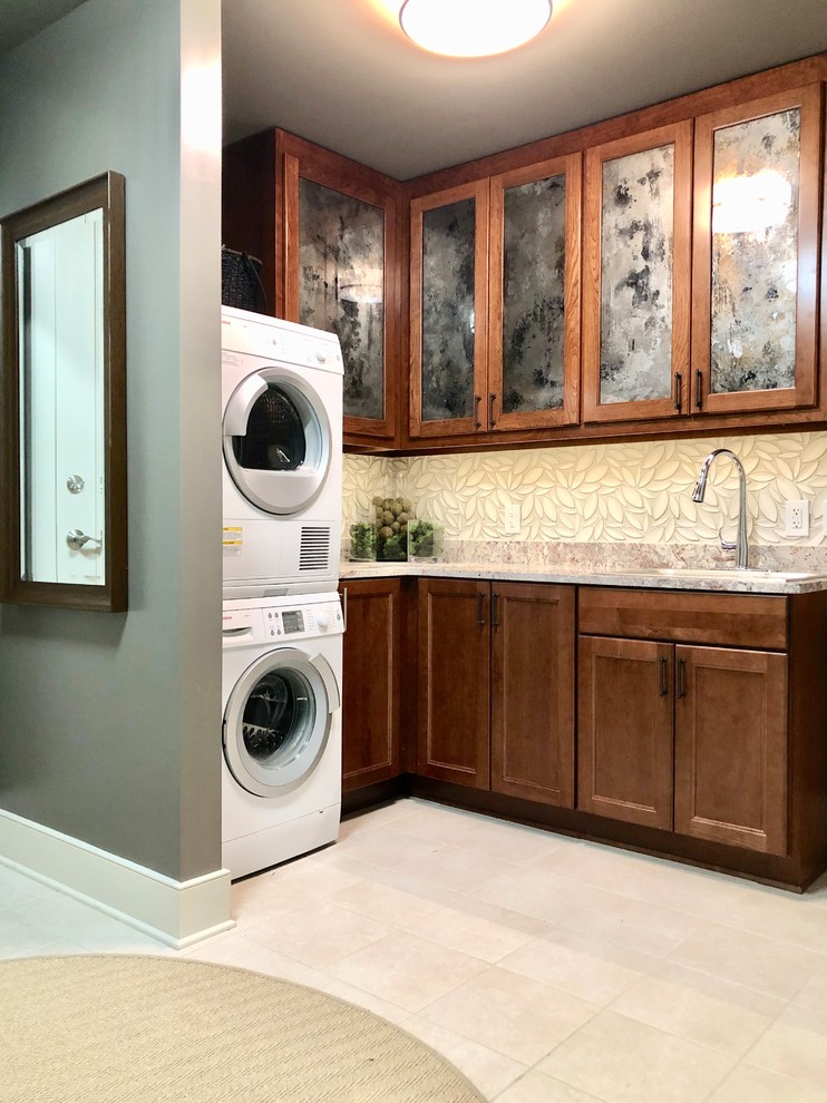 Photo of a large transitional galley dedicated laundry room in Atlanta with a farmhouse sink, shaker cabinets, brown cabinets, granite benchtops, grey walls, ceramic floors, a stacked washer and dryer, beige floor and multi-coloured benchtop.