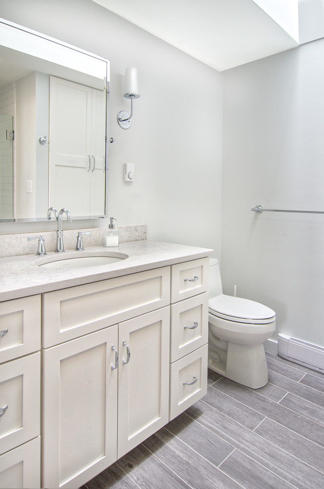 This is an example of a mid-sized beach style 3/4 bathroom in Portland Maine with shaker cabinets, yellow cabinets, an alcove shower, a one-piece toilet, white tile, subway tile, grey walls, porcelain floors, an undermount sink, grey floor, a hinged shower door and white benchtops.