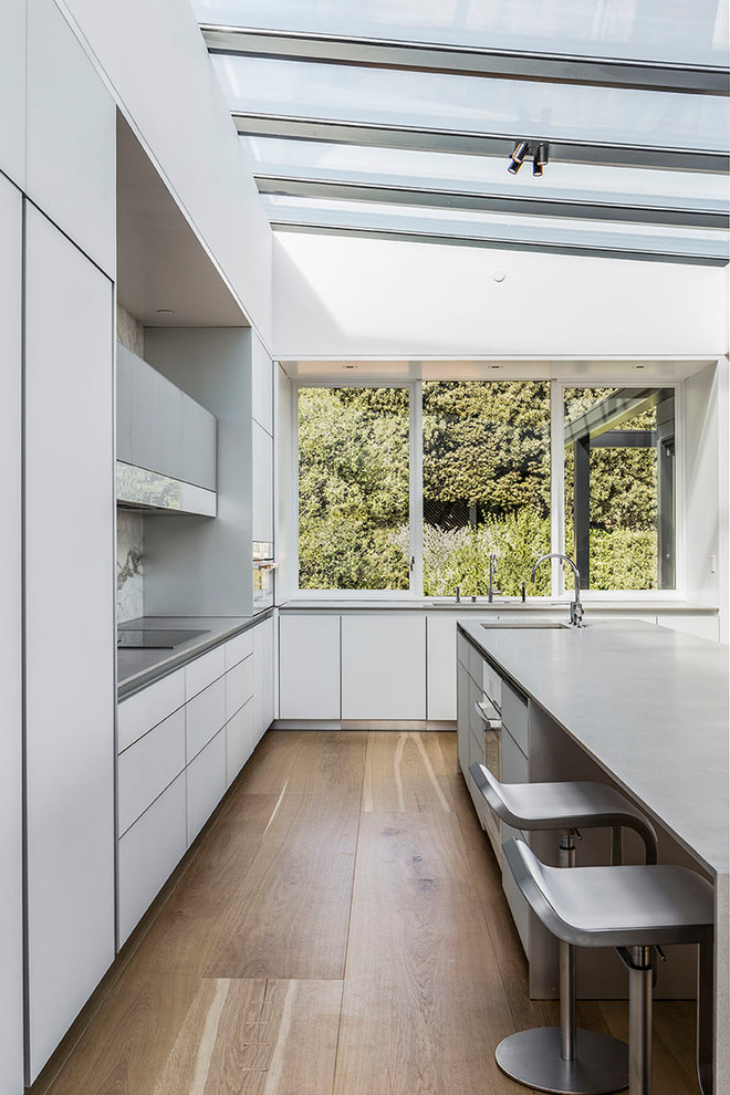 Photo of a large midcentury l-shaped eat-in kitchen in San Francisco with flat-panel cabinets, white cabinets, concrete benchtops, white splashback, marble splashback, white appliances, light hardwood floors and with island.