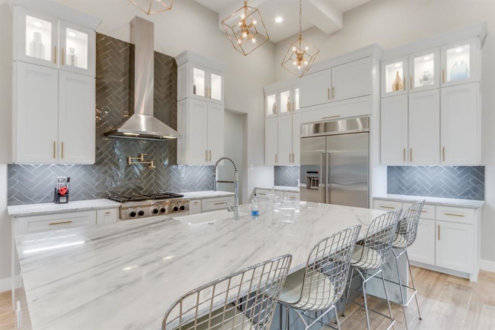 Photo of a large modern u-shaped open plan kitchen in Houston with a farmhouse sink, recessed-panel cabinets, white cabinets, marble benchtops, grey splashback, porcelain splashback, stainless steel appliances, light hardwood floors, with island, beige floor and grey benchtop.