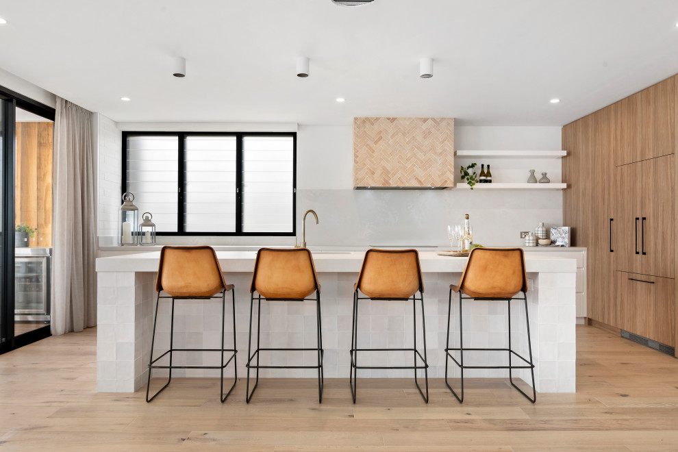 Inspiration for a beach style l-shaped kitchen in Sydney with flat-panel cabinets, medium wood cabinets, white splashback, panelled appliances, light hardwood floors, with island, beige floor and white benchtop.