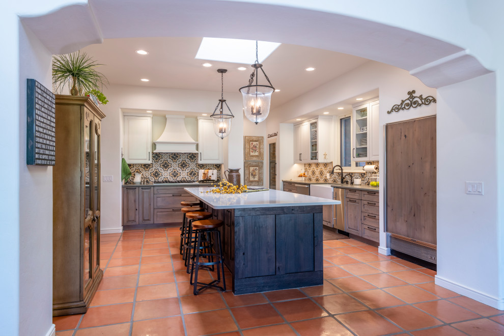 This is an example of a mid-sized u-shaped kitchen in Other with a farmhouse sink, raised-panel cabinets, distressed cabinets, quartz benchtops, multi-coloured splashback, cement tile splashback, stainless steel appliances, terra-cotta floors, with island, orange floor and white benchtop.