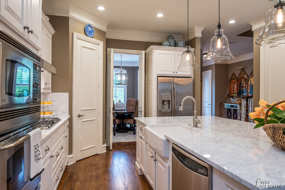 Mid-sized traditional l-shaped open plan kitchen in Dallas with a farmhouse sink, raised-panel cabinets, white cabinets, marble benchtops, white splashback, stone slab splashback, stainless steel appliances, medium hardwood floors and with island.