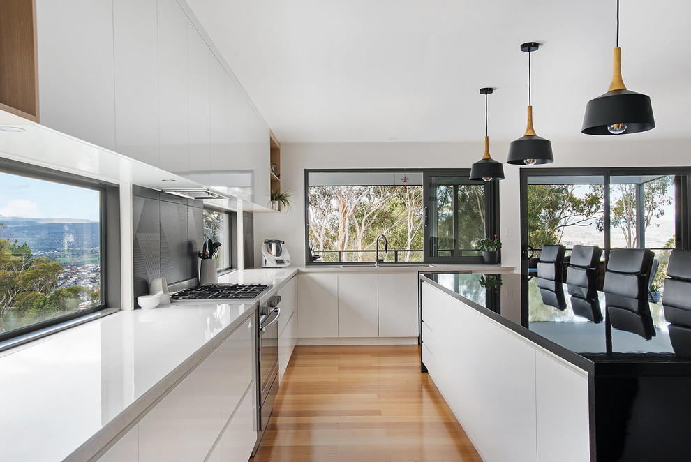 Mid-sized contemporary l-shaped eat-in kitchen in Hobart with a drop-in sink, white cabinets, laminate benchtops, laminate floors, with island, flat-panel cabinets, black splashback, black appliances, brown floor and white benchtop.