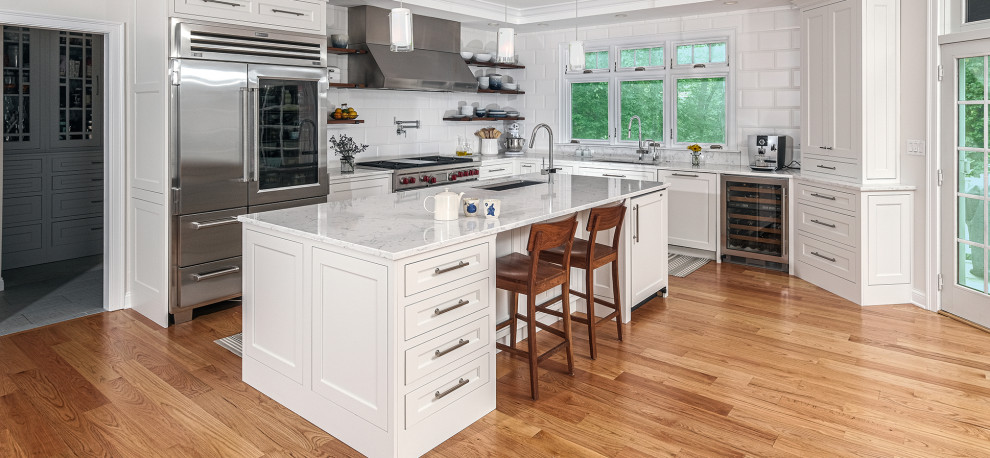 Photo of a mid-sized modern l-shaped eat-in kitchen in Boston with recessed-panel cabinets, white cabinets, marble benchtops, white splashback, stainless steel appliances, medium hardwood floors, with island, brown floor, white benchtop and an undermount sink.