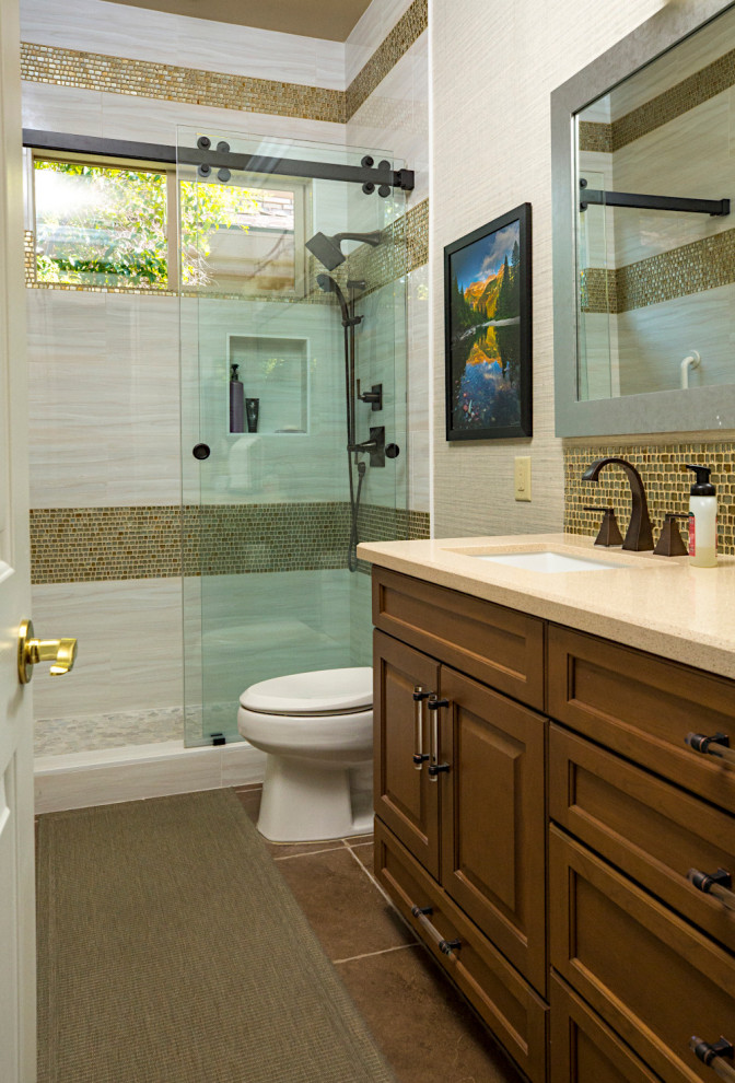 This is an example of a mid-sized traditional 3/4 bathroom in Las Vegas with raised-panel cabinets, brown cabinets, an alcove shower, a two-piece toilet, beige tile, glass sheet wall, green walls, porcelain floors, a drop-in sink, engineered quartz benchtops, brown floor, a sliding shower screen, beige benchtops, a single vanity, a built-in vanity, wallpaper and wallpaper.